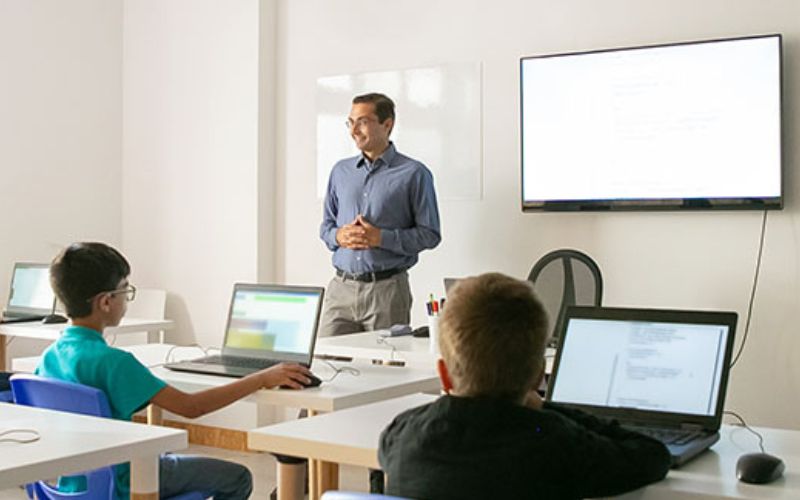 Teacher standing in front of class with a whiteboard behind him. Some students view the whiteboard screen on their laptops